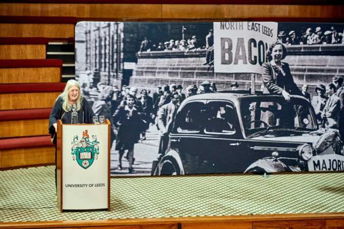 Professor Mary Beard delivering the Alice Bacon Lecture 2022. Professor Beard speaks at a lectern in front of a background photograph of Alice Bacon.
